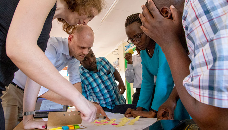 Group of people bending over a table. They are looking at a sheet of paper covered in Post-It notes.