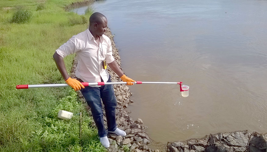 Man standing by water's edge. He is holding a cup with a water sample, which is attached to a short pole.