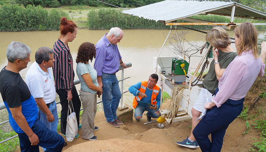 Group of people gathered round a water management device on the banks of a river in Peru. A man is showing them the workings of the device.