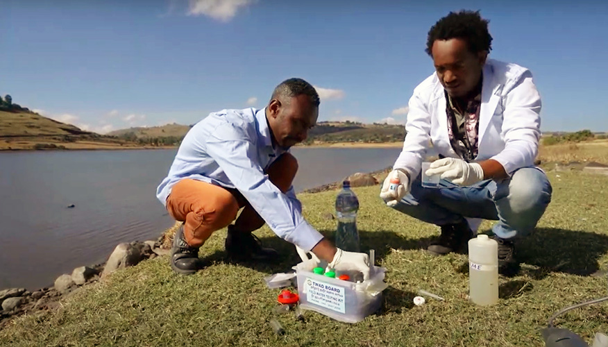 Water technicians are sat at the water's edge with a water testing kit, getting ready to take samples.