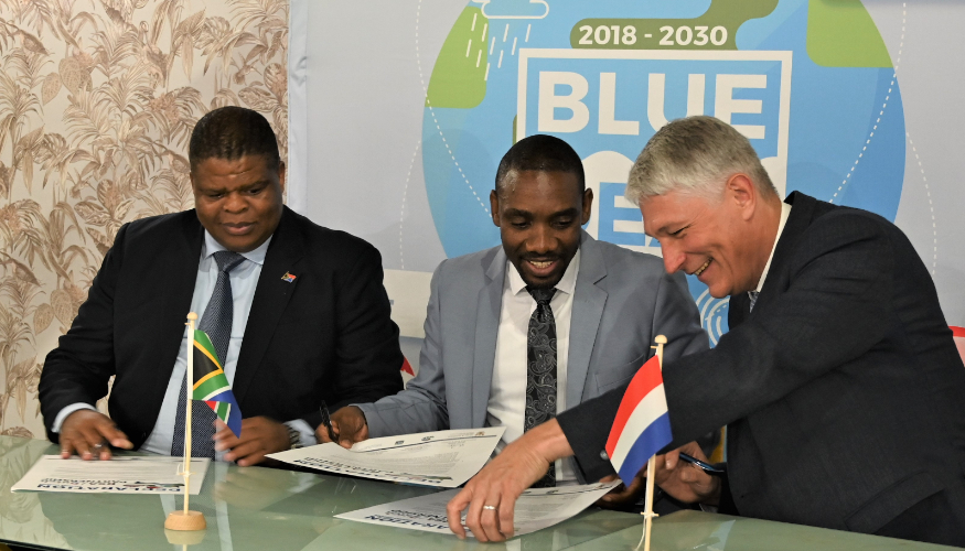 3 men sign a paper, with in the back a globe with the text Blue Deal on it.