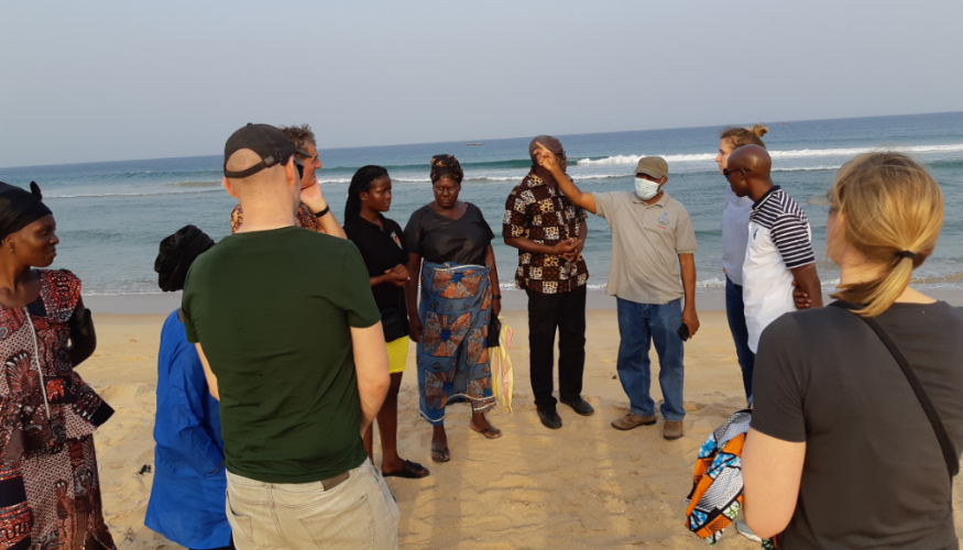 A group of people is standing on the beach where a person is explaining something.
