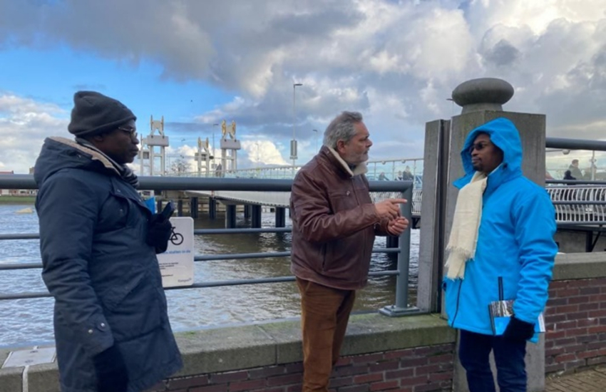 3 people are standing in front of a Dutch water work, 1 man is explaining something to the others.
