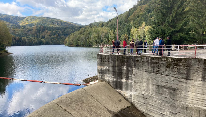 A group of people is standing next to a dam in Romania, with mountains in the background.