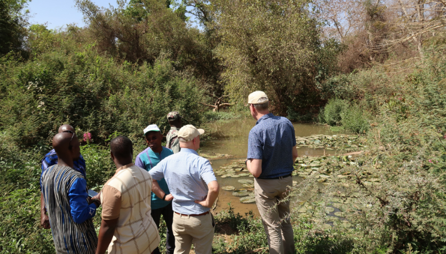 5 persons are standing in front of a small water, surrounded with trees.