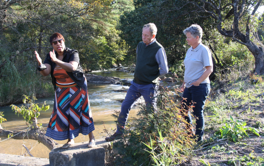 A woman is standing next to a river, explaining something to 2 other people. 