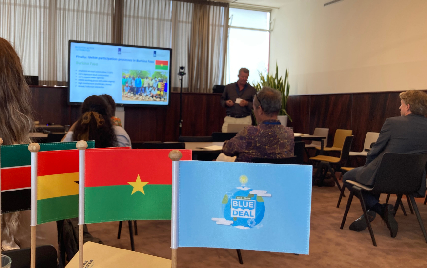 Photo of a small Blue Deal flag in front, with in the back somebody presenting a Powerpoint presentation.