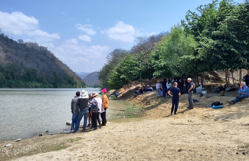 A group of people is standing next to a river, with mountains in the background.