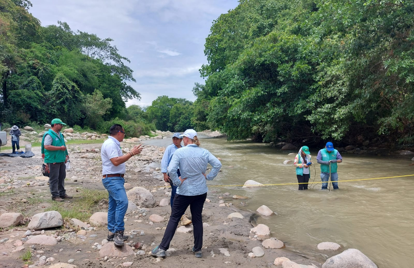 A group of people is standing on the bank of a small river. 2 persons are standing in the water for monitoring.