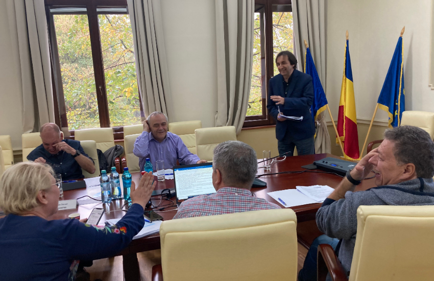 A group of people is sitting at a round table. One man is standing in front of 3 flags, with the Romanian flag in the middle.